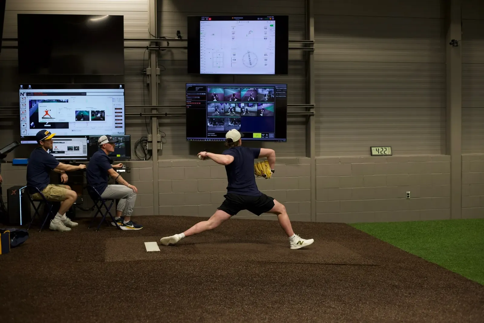 A pitcher mid-delivery on an indoor mound at BioPrecision’s lab while two staff review pitch-analysis software on wall-mounted screens.